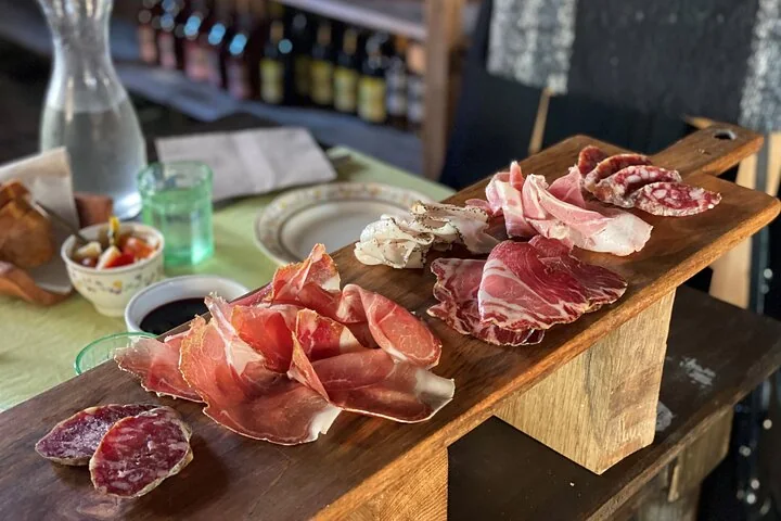 A wooden board of thinly sliced cured meats and salami on a table, with a carafe and glasses behind