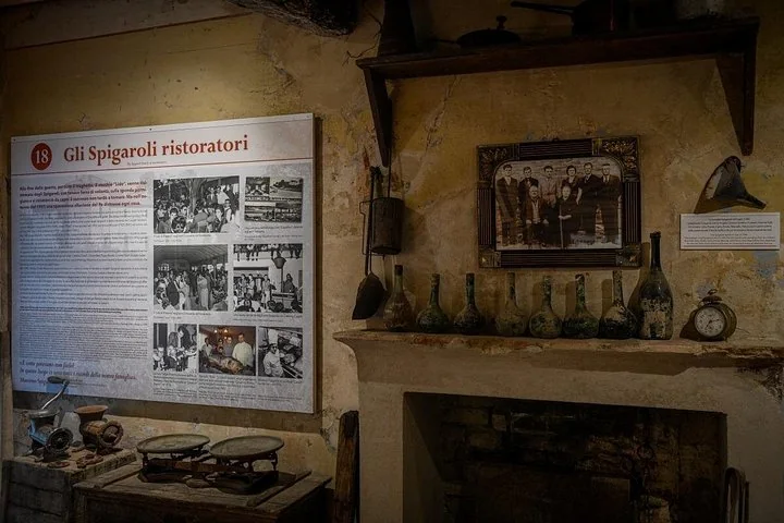 A museum room with a panel titled Gli Spigaroli ristoratori, old photographs, bottles on a mantelpiece and antique scales