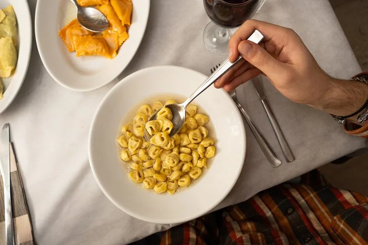 A hand spooning small stuffed pasta from a white bowl, with another plate of pasta and a glass of red wine on the table