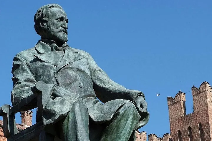 Una statua in bronzo di Giuseppe Verdi seduto contro il cielo azzurro, con dietro un castello di mattoni merlato